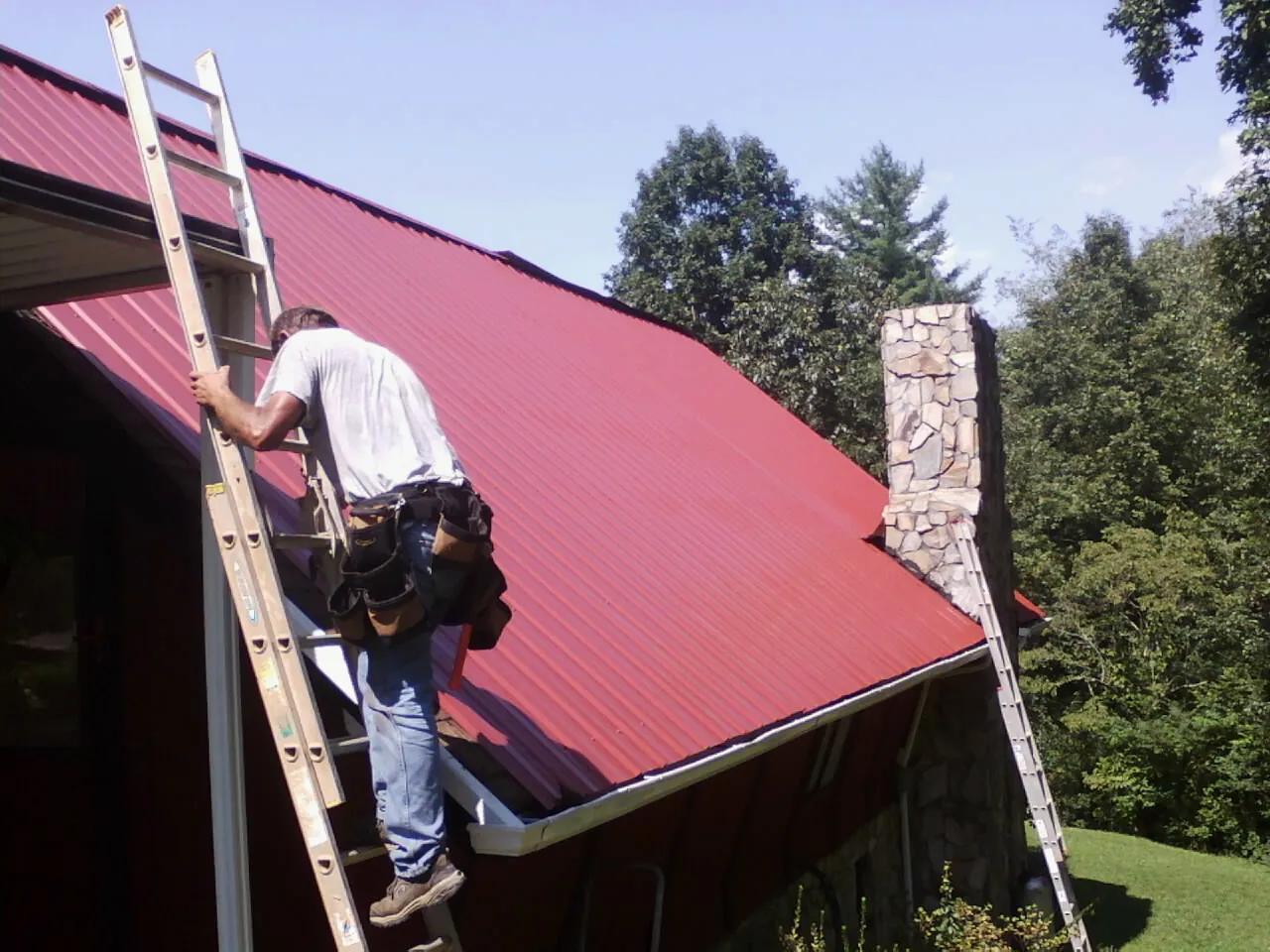 Roofer on a ladder beside a red metal roof and stone chimney — Soffit & Fascia Repair by Roofing Brothers in Oakland Gardens